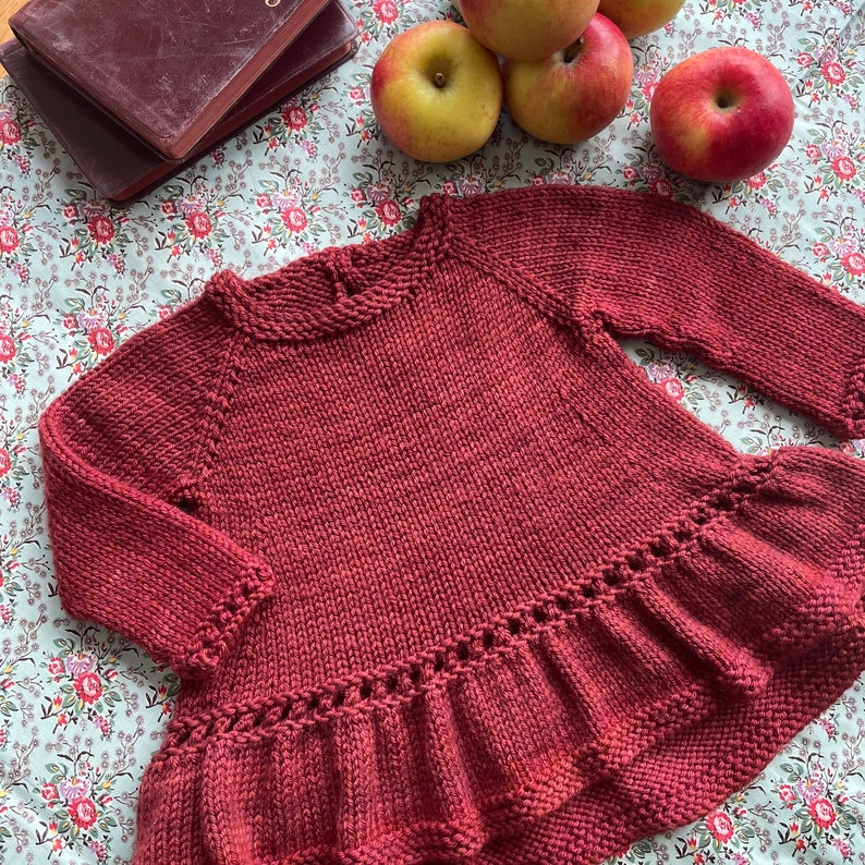 May include: A maroon knitted sweater with a ruffled hem. The sweater is made of a soft, textured yarn and has a relaxed fit.