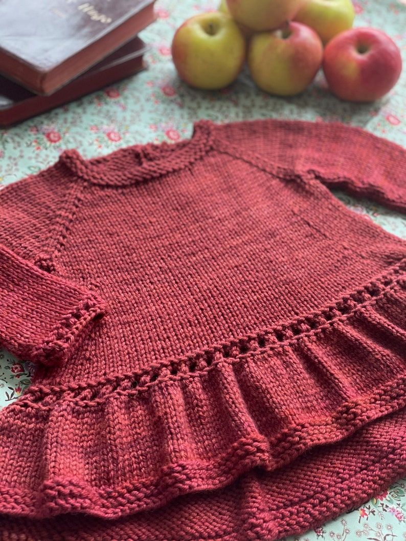 May include: A maroon knitted sweater with a ruffled hem. The sweater has a round neckline and long sleeves.