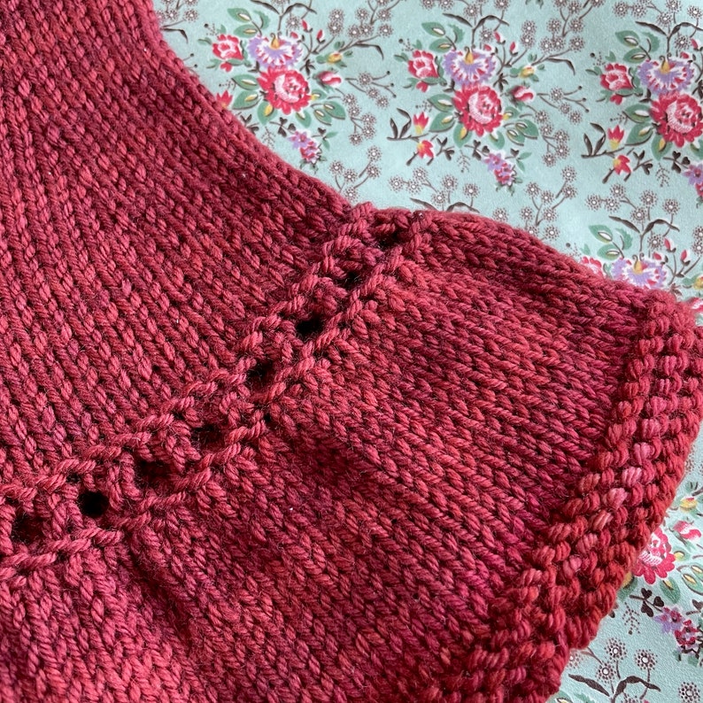 May include: Close-up of a deep red knitted fabric with a decorative stitch pattern along the edge.