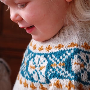 Pode incluir: Um close-up de uma criança vestindo um suéter de tricô branco com um padrão geométrico azul e dourado. A criança está olhando para baixo e sorrindo.