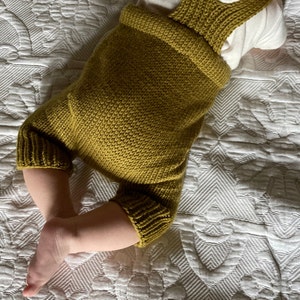 May include: A mustard yellow knitted baby romper with straps. The romper is made of a soft, textured fabric.