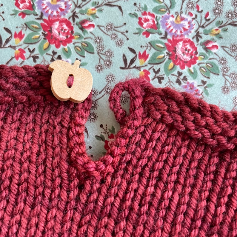 May include: A close-up of a burgundy knitted fabric with a wooden button shaped like an apple. The fabric has a textured, woven appearance.