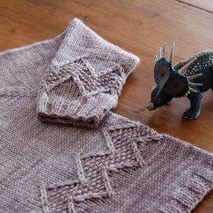 Puede incluir: Primer plano de un suéter tejido con un patrón de punto de cable en color morado claro. El suéter tiene un juguete de dinosaurio a su lado.