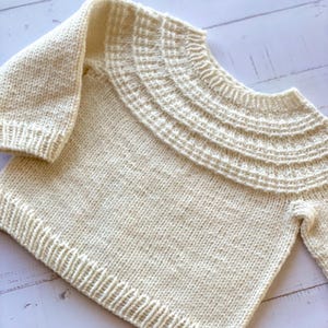 Könnte beinhalten: Cremefarbener Strickpullover mit breitem Rippenkragen und langen Ärmeln. Der Pullover hat ein strukturiertes Strickmuster und einen gerippten Saum. Das Design ist schlicht und gemütlich, passend für kühlere Temperaturen.