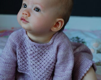 May include: A baby wearing a light purple knitted sweater with a textured pattern. The baby is sitting on a colorful rug and looking up.