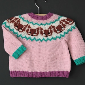 Könnte beinhalten: Ein rosa gestrickter Pullover mit einem grün-weißen Muster am Joch, das braune Füchse zeigt. Der Pullover hat einen violetten Saum am Hals und am unteren Rand.