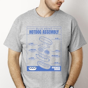Puede incluir: Una camiseta gris con un plano azul de montaje de perrito caliente. El diagrama muestra los componentes: pan, queso, carne picada, pepinillos, salsa de mostaza y salchicha. El texto dice "Blue Print Hotdog Assembly Schematic Diagram."