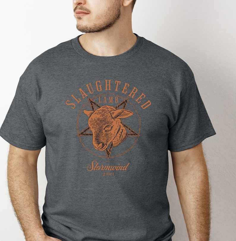 May include: Dark gray t-shirt featuring a distressed orange graphic. The graphic includes the words "Slaughtered Lamb" above a lamb's head within a star, with "Stormwind" and "2001" below. The shirt has a crew neck.