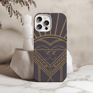 May include: A phone case with a dark gray background and a gold art deco design. The design includes a heart shape, sunburst rays, and the number "33" in a triangle. The case has a camera cutout.