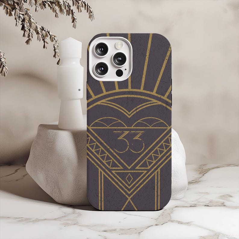 May include: A phone case with a dark gray background and a gold art deco design. The design includes a heart shape, sunburst rays, and the number "33" in a triangle. The case has a camera cutout.