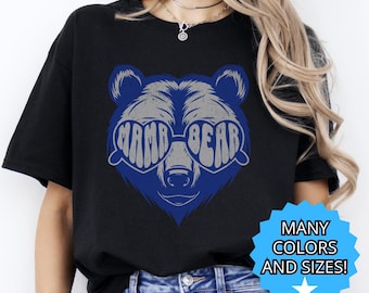 以下が含まれることがあります： 黒いTシャツ。青いクマの顔のデザインで、サングラスに「MAMA BEAR」というグレーの文字が書かれています。デザインは青いアウトラインで囲まれています。青いスターバーストグラフィックには「MANY COLORS AND SIZES！」と書かれています。
