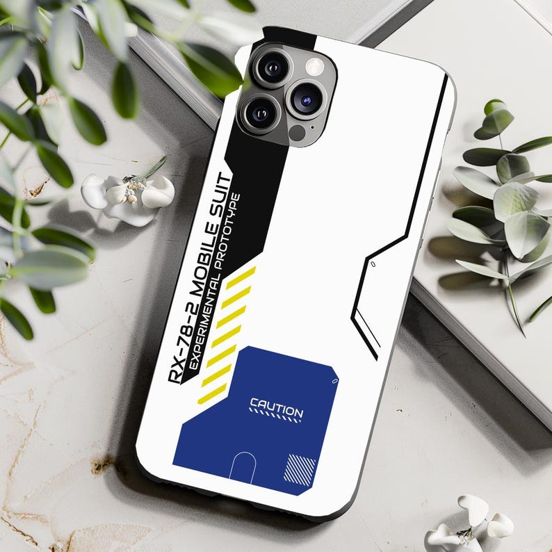 May include: White phone case with a black and yellow design. The case has the text "RX-78-2 MOBILE SUIT EXPERIMENTAL PROTOTYPE" and "CAUTION" printed on it.