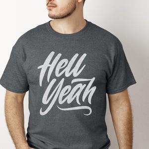 Può includere: T-shirt grigio scuro con la scritta bianca "Hell Yeah" in corsivo. La maglietta ha un girocollo e maniche corte. Il testo è centrato sulla parte anteriore.