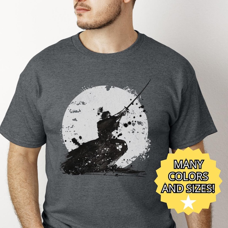 K&ouml;nnte beinhalten: Dunkelgraues T-Shirt mit einer Schwarz-Wei&szlig;-Illustration eines Samurai mit Schwert vor einem wei&szlig;en Mond. Das Shirt hat den Text "MANY COLORS AND SIZES!" auf einem gelben Stern.