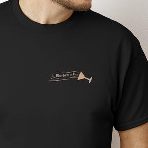 Może przedstawiać: Czarny T-shirt z brązową grafiką kieliszka do martini i tekstem "Blurberry Bar Grand Tree"