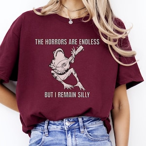 K&ouml;nnte beinhalten: Ein maroonfarbenes T-Shirt mit einem Grafikdruck einer Gitarre spielenden Frosch. Der Text auf dem Shirt lautet "The horrors are endless but I remain silly."