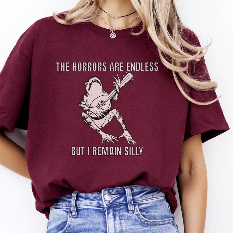 K&ouml;nnte beinhalten: Ein maroonfarbenes T-Shirt mit einem Grafikdruck einer Gitarre spielenden Frosch. Der Text auf dem Shirt lautet "The horrors are endless but I remain silly."