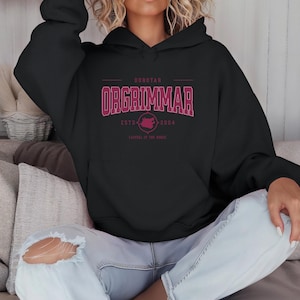 Puede incluir: Sudadera con capucha negra con bolsillo delantero y capucha. La parte delantera de la sudadera tiene el texto "ORGRIMMAR" en color granate, con texto adicional y un gráfico debajo. La sudadera es usada por una persona con jeans rotos.