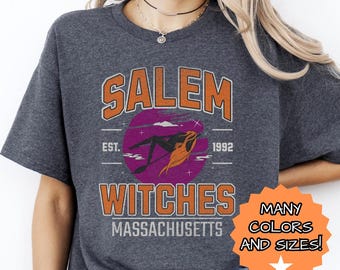 Puede incluir: Una camiseta gris oscuro con un diseño gráfico de estilo vintage que dice "Salem Witches Massachusetts Est. 1992" con una silueta de una bruja volando en una escoba. El gráfico es en naranja, morado y blanco.