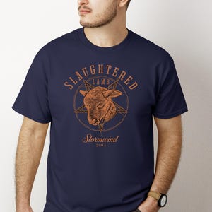May include: Navy blue t-shirt with a distressed orange graphic. The graphic features a lamb's head inside a pentagram, with the words "SLaughtered Lamb" above and "Stormwind 2001" below. The shirt is a crew neck.