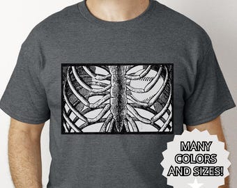 May include: Dark gray t-shirt featuring a black and white illustration of a rib cage with a baseball inside. The shirt has the text "MANY COLORS AND SIZES!" on a white starburst graphic.