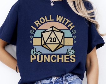 May include: Navy blue t-shirt with a graphic design featuring a D20 die and the text "I ROLL WITH THE PUNCHES". The design includes a retro color scheme with a beige, teal, and orange striped background. The shirt is a casual wear item.