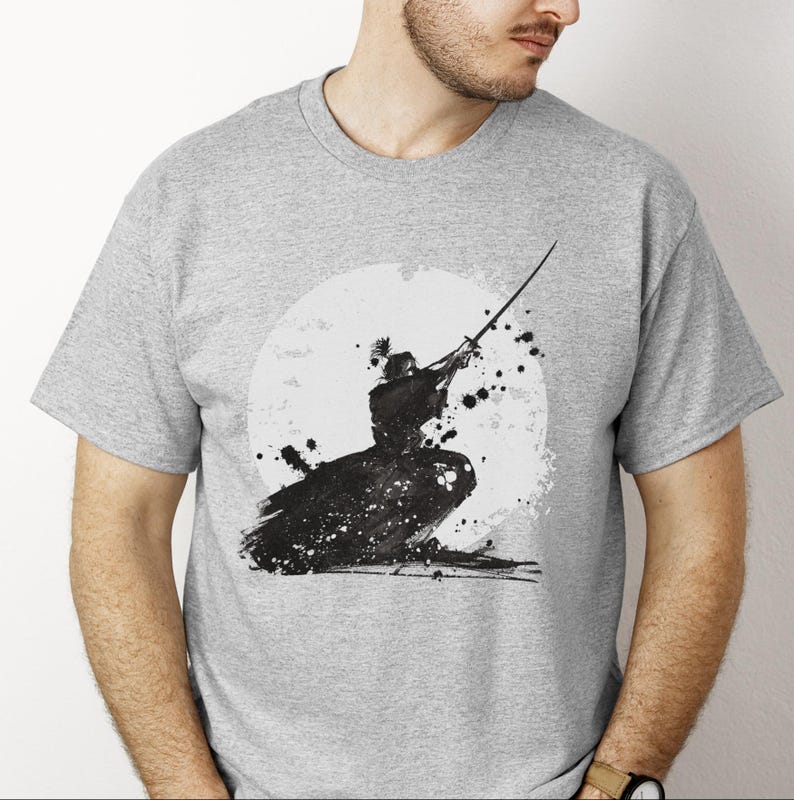 K&ouml;nnte beinhalten: Hellgraues T-Shirt mit einer Schwarz-Wei&szlig;-Tuscheillustration eines Samurai-Kriegers in Aktion. Das Design umfasst einen wei&szlig;en kreisf&ouml;rmigen Hintergrund und schwarze Farbspritzer, die einen dynamischen und k&uuml;nstlerischen Look erzeugen.