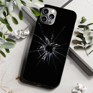 May include: Black phone case with a cracked screen design. The case features a glossy finish and a realistic shattered glass effect, centered around the Apple logo. The case is shown on a marble surface with greenery.