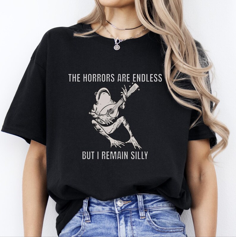 K&ouml;nnte beinhalten: Schwarzes T-Shirt mit einer wei&szlig;en Grafik eines Frosches, der Banjo spielt. Der Text "The horrors are endless but I remain silly" ist auf das T-Shirt gedruckt.