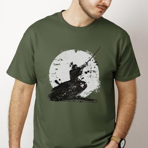 K&ouml;nnte beinhalten: Olivgr&uuml;nes T-Shirt mit einer Schwarz-Wei&szlig;-Illustration eines Samurai-Kriegers mit Schwert, vor einem wei&szlig;en, gesprenkelten Mond. Das Design hat einen Pinselstrich-Stil, der ihm ein dynamisches und k&uuml;nstlerisches Aussehen verleiht.