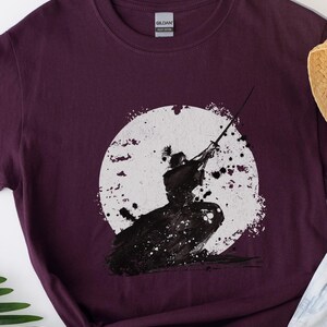 K&ouml;nnte beinhalten: Burgunderfarbenes T-Shirt mit einer Schwarz-Wei&szlig;-Illustration eines Samurai vor einem wei&szlig;en Kreis. Der Samurai wird in einer dynamischen Pose mit einem Schwert dargestellt. Das Design hat eine aquarellartige &Auml;sthetik.