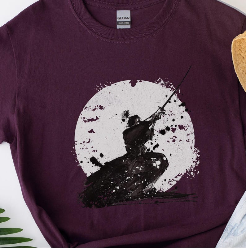 K&ouml;nnte beinhalten: Burgunderfarbenes T-Shirt mit einer Schwarz-Wei&szlig;-Illustration eines Samurai vor einem wei&szlig;en Kreis. Der Samurai wird in einer dynamischen Pose mit einem Schwert dargestellt. Das Design hat eine aquarellartige &Auml;sthetik.