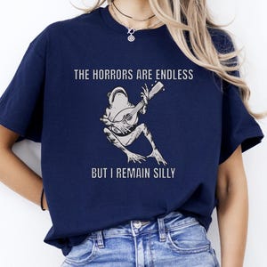 K&ouml;nnte beinhalten: Ein marineblaues T-Shirt mit einer wei&szlig;en Grafik eines Frosches, der Banjo spielt. Der Text "The horrors are endless but I remain silly" ist auf das T-Shirt gedruckt.