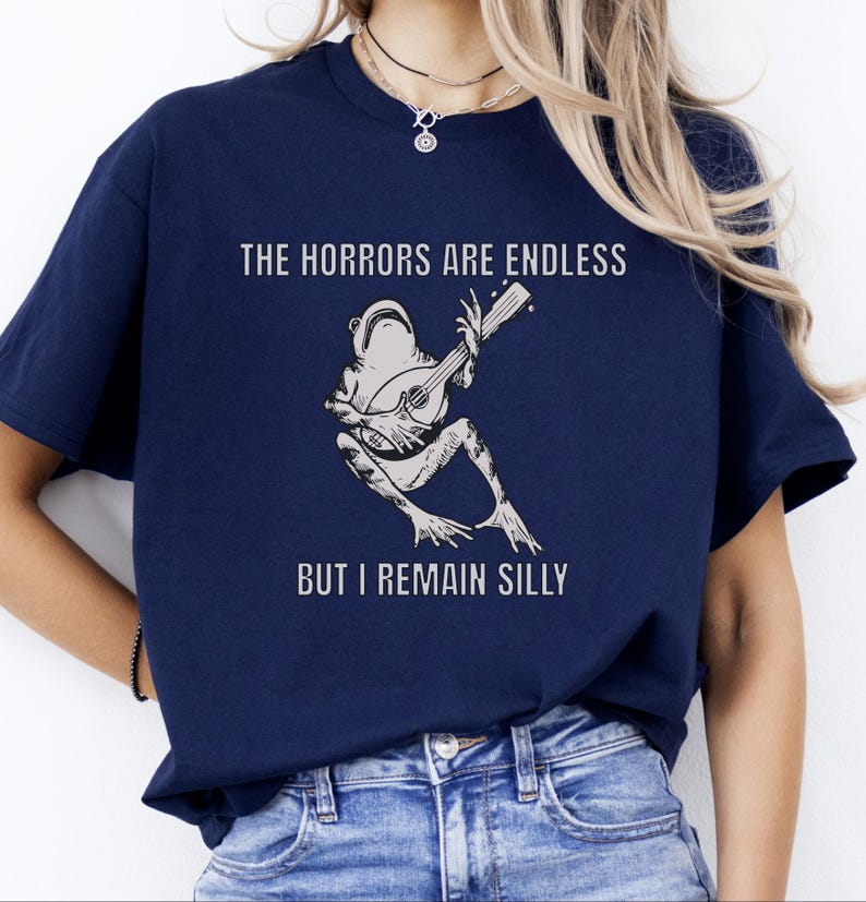 K&ouml;nnte beinhalten: Ein marineblaues T-Shirt mit einer wei&szlig;en Grafik eines Frosches, der Banjo spielt. Der Text "The horrors are endless but I remain silly" ist auf das T-Shirt gedruckt.