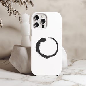 Puede incluir: Funda de teléfono blanca con un diseño de círculo negro. La funda tiene un recorte para el objetivo de la cámara y un fondo blanco.