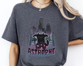 Puede incluir: Camiseta gris oscuro con un gráfico de gato negro y el texto "CAT ASTROPHE". El gato está representado frente a un horizonte urbano con acentos morados y azules. La camiseta tiene mangas cortas y cuello redondo.