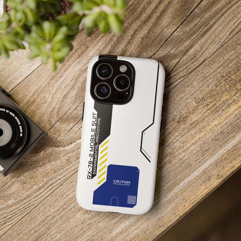 May include: White phone case with a black and gray design. The case has a blue and yellow graphic with the text "RX-78-2 MOBILE SUIT EXPERIMENTAL PROTOTYPE" and "CAUTION".