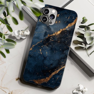 May include: A smartphone case with a dark blue marble design and gold veining. The case has a glossy finish and a camera cutout. The case is lying on a white surface with greenery.