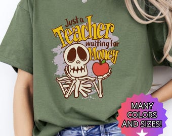 May include: Green t-shirt with a graphic featuring a skeleton holding a red apple. The text reads "Just a Teacher waiting for Money" in yellow and white. A multi-colored starburst graphic at the bottom says "MANY COLORS AND SIZES!"