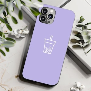 May include: A light purple phone case featuring a white outline of a bubble tea drink. The case is made of a smooth material and has a camera cutout. The phone case is resting on a white surface with greenery.