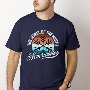 May include: Navy blue t-shirt featuring a retro design with the text "THE JEWEL OF THE NORTH" and "Neverwinter". The design includes a snowflake graphic with orange, pink, blue, and white colors.