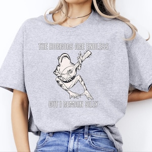 K&ouml;nnte beinhalten: Ein graues T-Shirt mit dem Text "The horrors are endless but I remain silly" und einer Schwarzwei&szlig;illustration eines Frosches, der Gitarre spielt.