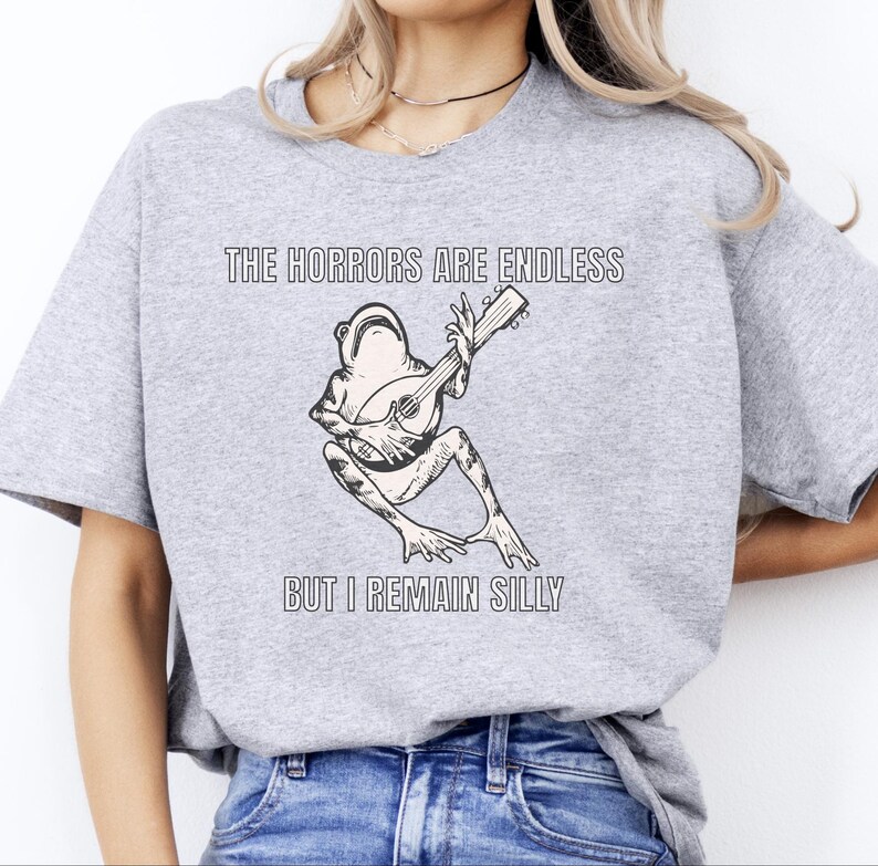 K&ouml;nnte beinhalten: Ein graues T-Shirt mit dem Text "The horrors are endless but I remain silly" und einer Schwarzwei&szlig;illustration eines Frosches, der Gitarre spielt.