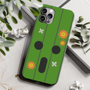 Op de afbeelding: Groene telefoonhoes met een cartoon gezicht en bloemmotieven. De hoes heeft een zwarte camera lens en een wit bloemenontwerp.