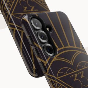 May include: Black phone case with a gold art deco design. The case features a geometric pattern with sunburst and zigzag elements. The case has a glossy finish and a camera module with three lenses.
