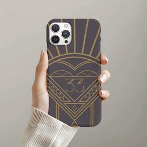 May include: A dark gray phone case with a gold art deco design. The design includes a heart shape, sunburst rays, and the number "33" in a triangle. The case is held in a hand.