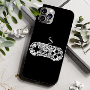May include: Black phone case with a white, detailed illustration of a video game controller. The controller design is made up of various gaming-related icons. The phone case is shown on a white surface with green foliage.