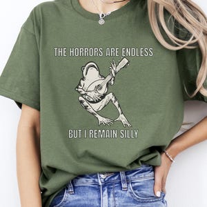 K&ouml;nnte beinhalten: Ein gr&uuml;nes T-Shirt mit einer Grafik einer Gitarre spielenden Frosch. Der Text auf dem Shirt lautet "The horrors are endless but I remain silly."