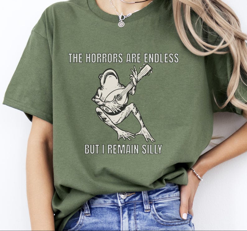 K&ouml;nnte beinhalten: Ein gr&uuml;nes T-Shirt mit einer Grafik einer Gitarre spielenden Frosch. Der Text auf dem Shirt lautet "The horrors are endless but I remain silly."