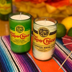 May include: Two glass candles with white wax, one green and one clear, feature labels reading "Topo Chico" and "Twist of Lime." The candles sit on a colorful striped textile, with limes, a lemon, and a small red umbrella in the background.